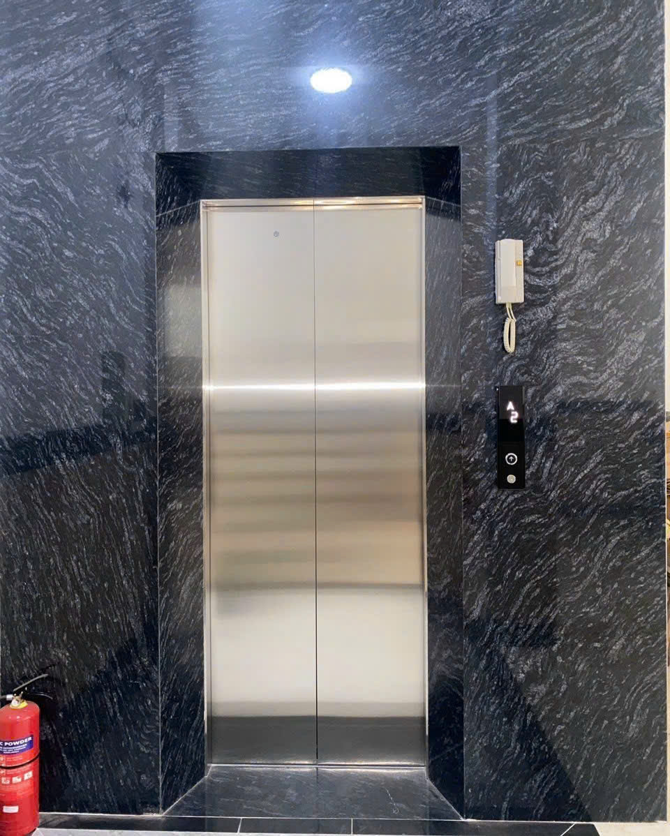 Thang máy cửa inox đóng kín trong sảnh ốp đá đen, bên cạnh có bảng gọi tầng và bình chữa cháy